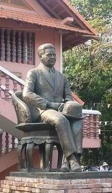 A cast-iron statue of a man in a suit with a hat in his lap, seated upon a chair also of cast iron; the wall and roof of a building is visible in the background.