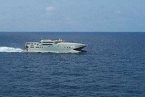 A warship-grey catamaran travelling at speed (from right to left) on the open sea