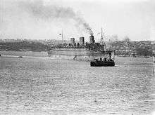 Black and white photo of a large passenger ship in an enclosed harbour. A smaller ship is visible in front of the passenger ship.