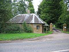 Small building to the left of gateposts surrounded by trees