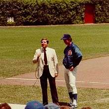 Two men standing on a grass-and-dirt field; the man on the right is wearing a blazer and slacks, while the man on the left—the subject of the image—is wearing white pinstriped baseball pants, a blue nylon warm-up jacket, and a blue baseball cap.