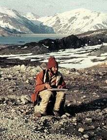 Man wearing a coat and hat and holding a pad of paper sits on a rock , with a lake and several mountains visible in the background