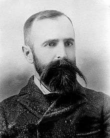 Portrait shot of a man perhaps around 40 (Wilson).  He is dressed in archaic clothing, with a checked suit jacket close around his neck.  His hair is cut close, but he wears a beard and an impressive mustache, with straight hair from it extending well to each side of the face.