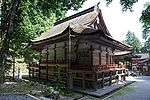 Three-quarter view of a proportionally tall wooden building with a veranda with red hand rail. The veranda is higher at the center of the back side of the building. The corner of the eaves appear cut off.