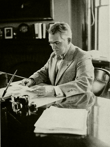 Half-length portrait of a bespectacled man in a suit and bow-tie sitting at a desk, he is looking intently at a piece of paper on top of a small stack of others. Another stack sits in front of him to his left. A clock sits on a mantle behind him with a small picture frame and several books, to its right is a window which fills the room with soft light.