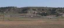 Fenced area with wind vane and antennas near base of rocky hills