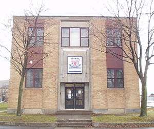  A two story brick building with four large windows evenly spaced, and a large logo in between them, like the number five on a die, two leafless trees flank the building's entrance.