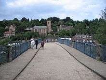 A view looking North along the roadway across the bridge, the curve  of its arch obscuring much of the town of Ironbridge behind. The town rises up the steep gorge side beyond with the Tontine Hotel and the Church of St Luke directly ahead.