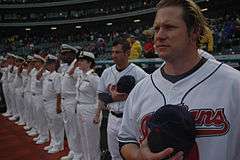 A man with wavy swept-back hair wearing a white baseball jersey and holding a baseball cap over his heart standing at the end of a row of like-dressed men