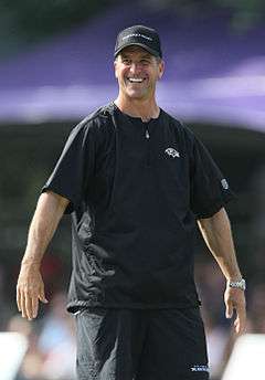 Color photograph of tall, smiling man (John Harbaugh) dressed in black sport shirt, black practice shorts, and black baseball cap.