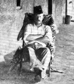 A middle-aged, moustachioed gentleman sits with his arms folded outside a house.