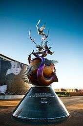 A colour photograph of a large metal monument with a conical base supporting a globe that is wrapped in contorted musical instruments. In the background is a blue sky.