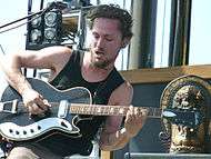 Butler is sitting strumming the strings with his right hand while his left hand is on the fret board. His hair is relatively short and he is bearded. A microphone is above and to his right with other stage equipment behind his right shoulder. To his left is an oriental statue.