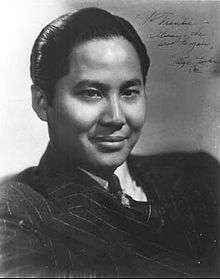 Head and shoulders publicity shot of a young, smiling man, short hair slicked back and wearing a suit and tie.