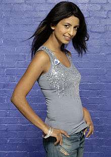 A woman with black hair wearing a sparkly, light-blue shirt and blue jeans standing in front of a shiny, blue-bricked wall and looking at the viewer