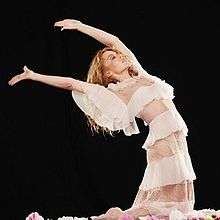 Minogue kneeling in a white dress, leaning back with her hands held up.