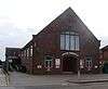 Three-quarter view of a dark brick building with lighter brick dressings around the arched entrance.  "LANCING TABERNACLE" is written in white letters above this and below a wide, tall, slightly arched window with brown mullions.  Evenly-spaced rectangular windows extend down the longer side.  A brick-paved area is in front.