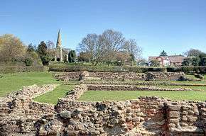 Low reddish-coloured stone walls, the construction of which suggests antiquity, lie in a grassy area backed by the outline of a modern settlement including a church with a spire. The trees lack leaves and the sky is a clear blue.