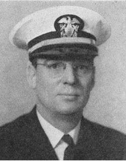 Head of a middle-aged white man wearing a white peaked cap, wire frame glasses, and a dark jacket over a shirt and tie.