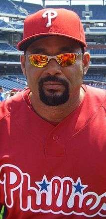 An olive-skinned man with a dark goatee and mustache wearing dark sunglasses, a red baseball cap with a white "P" on the face, and a red baseball jersey with "Phillies" in white and red script across the chest