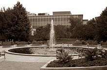 a large, flat-roofed building, partially obscured by trees, with a fountain in a circular pond in the foreground