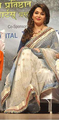 Front profile of an Indian woman, sitting on a chair, wearing a white glittering sari. She has a hint of smile on her face and her dark-brown hair falls in curls on her right shoulders.