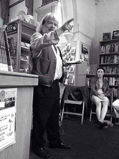 photo of a rotund bearded jolly man pointing to the audience with one hand while reading from the book of his poetry with his other.