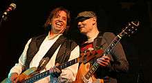 Mark Tulin—a middle-aged Caucasian male with long brown hair wearing a white shirt and black vest—plays bass guitar and smiles while Billy Corgan—a middle-aged Caucasian male wearing a dark green hat and red-and-black striped shirt with a brown jacket—plays electric guitar to his left.