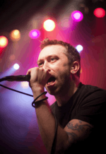 A man holding a mircophone sings in front of a red and blue background.