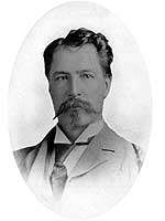 Head and shoulders black-and-white photo of a man in his twenties, wearing a late Victorian style suit and tie, with slightly unkempt wavy dark hair and a lighter-coloured goatee. He has a determined look on his face.