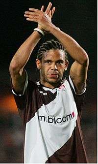 African man in a diagonally striped brown-and-white jersey with a sponsor logo across the chest. His hair is cornrowed, and his arms are crossed above his head.