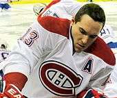 Close up view of a hockey player as he stretches prior to a game. He has short black hair and is wearing a red, white and blue uniform with a stylized "CH" logo on his chest and the letter "A" denoting his position as alternate captain.