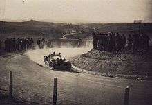 An old sports car drives on unsealed roads as people stand on hills around the road and watch