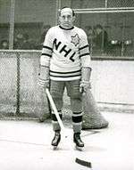 An ice hockey player stands on an ice rink. He is wearing a sweater that has the letters "NHL" in a downward diagonal with a large star on his left shoulder.