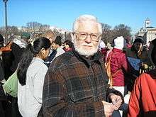 Picture of a gray-haired man wearing glasses and a brown plaid flannel shirt; behind him is a crowd of people