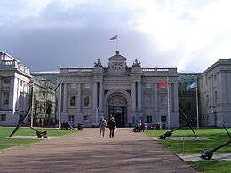  Facade of an ornate 18th century building, with tall stone columns, a wide arched entrance, balustraded roof and a central pediment from which a flag is flying. The building is approached by a wide path flanked by lawns.