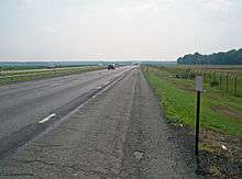 An expressway, seen from the shoulder of its righthand lanes, continues straight ahead to the center of the image. Either side is fenced off; the surrounding area is mostly marsh with a wooded area visible at right center