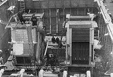 A cavernous steel room, with two large rectangular boilers inside; a number of men are working on the machines.