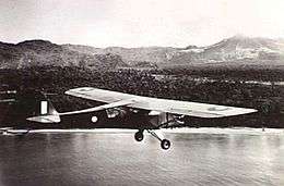 Single-engined high-wing light plane flying over water, with jungle terrain in background