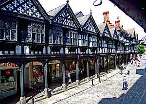 A street containing a range of two-storey shops on the left seen from a slightly elevated position.  The lower storey contains modern shop fronts behind an arcade; the upper storey is timber-framed and contains a variety of windows and decorated gables.