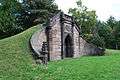 A small knoll rises from the left and his held back by a brown, gothic stone wall with gothic entrance. A shield is carved above the entrance and contains, among other words, the letters VAIL.