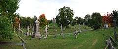 A panoramic view of the cemetery; a dirt road pass in front of the view from right to left. At center is a grey church-like structure, surrounded on all sides by various sized grave stones.