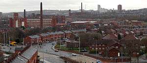 A view of a busy townscape, in which the land is urban and the scene is banal. The weather is overcast and the cloudy sky appears light-grey. In the foreground is a dark-grey tarmac dual-carriageway road, which sweeps up the middle of the photograph to its centrepoint. All around the road are two-storey red-brick houses. On the left-side of the midground are three large red-brick factories of around five-storeys high. On the horizon is a towerblock.