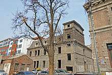 A tan building similar to the main house with a tower on the right. In front of it is a large bare tree. To the lower right is a small dilapidated brick building with a gabled roof.