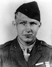 Head of a young man in military uniform with a quizzical look on his face. His garrison cap is tilted off to one side and a tuft of short hair sticks out from the other.