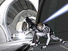 A horizontally rectangular video game screenshot that is a digital representation of a space station. A woman in a white and grey suit holding a large black gun ducks underneath a blue beam of light.