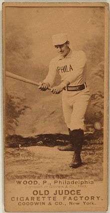 A sepia-toned baseball card image of a man wearing an old-style white baseball uniform and pillbox cap holding a baseball bat extended with both hands