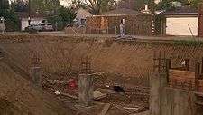 A woman stands in the far background in front of a house, overlooking a large dir put filled with debries, planks of wood and unfinished concrete structures.