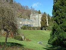 A large stone house in the middle distance, framed by trees in the foreground, and with a sloping lawn in front.  The left part of the house has two storeys, while the right section is much taller, in four storeys, surmounted by battlements.