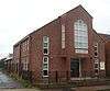 A modern building in two shades of red brick, behind a low black fence. White-framed three-paned windows line the side wall and flank the entrance—a double door sheltered below a protruding section supported on brick columns.  This has a plaque reading "QUEEN STREET CHURCH CENTRE" and a pentagonal window.